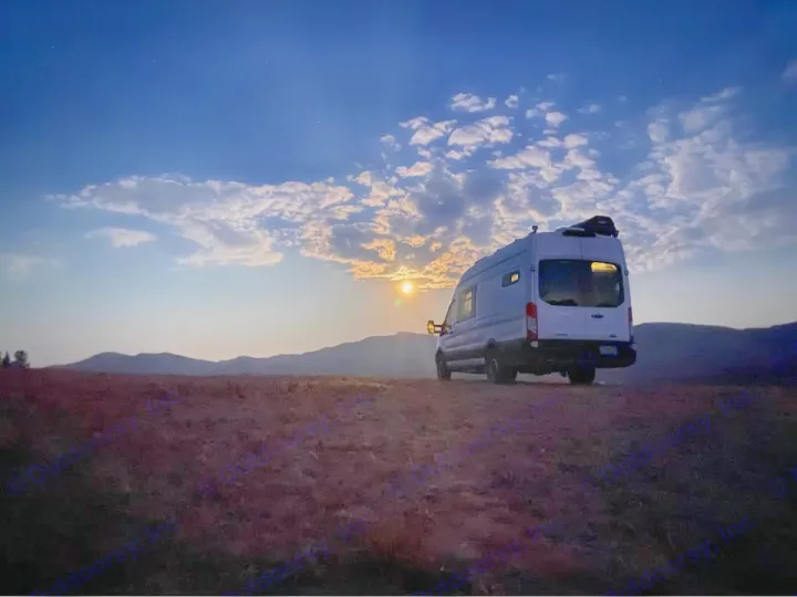 White camper van (RV rental) at sunset/moonrise on a hill overlooking mountains, under a cloudy sky. Serene view.. Ford Transit 2018