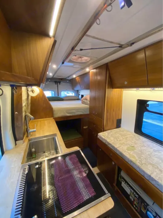 Modern RV rental camper van interior. Features a wood-finished kitchen, raised bed, and clever storage solutions. Well-lit and cozy.. Ford Transit 2018