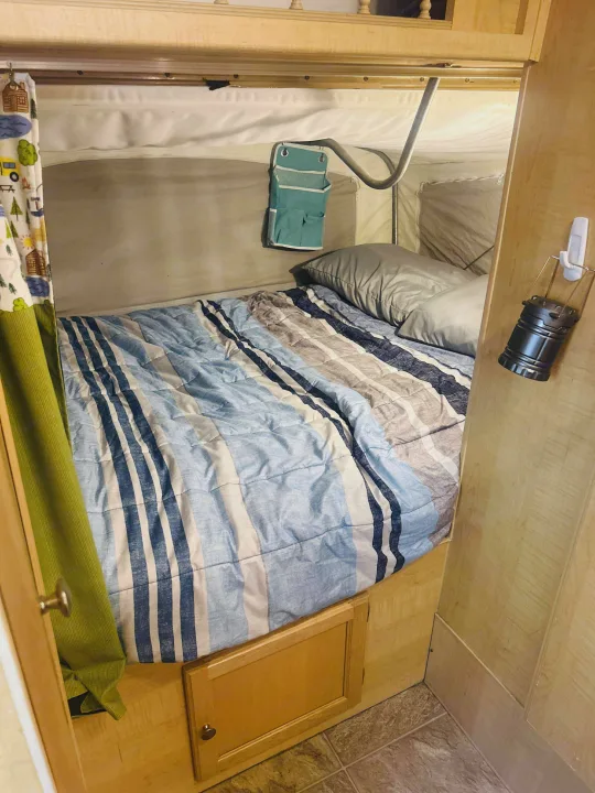 Cozy bed inside an RV rental with striped bedding, a camping-themed curtain, storage, and a lantern.. Forest River M-17 2005