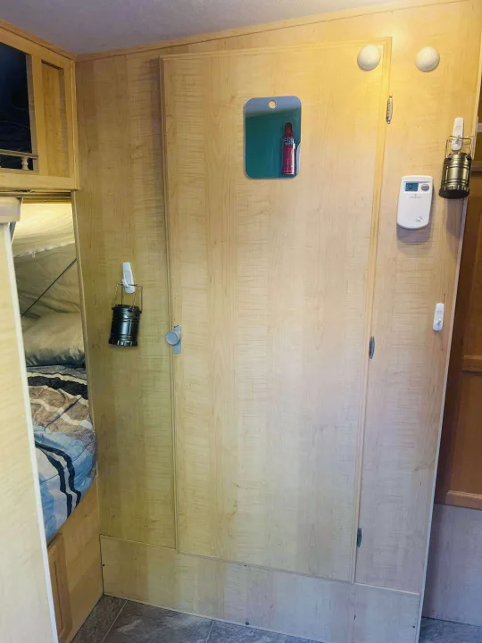 Inside an rv rental: light wood cabinetry with a mirror, thermostat, and hanging lantern. A comfy bed is glimpsed within.. Forest River M-17 2005