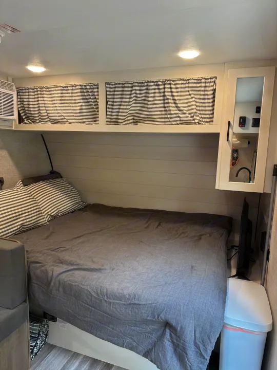Cozy RV rental bedroom featuring a bed with gray linens, striped pillows, overhead storage, AC, and a TV.. Jayco SLX Sport 2025