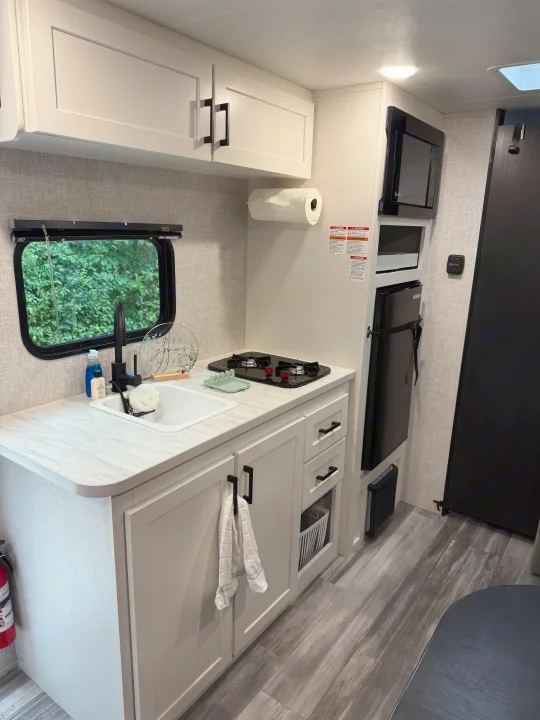 Compact and modern RV rental kitchen: white cabinets, sink, two-burner stove, microwave, and fridge. Your mobile cooking space.. Jayco SLX Sport 2025