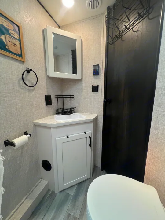 Inside an RV rental: a compact bathroom with a white corner vanity, sink, mirrored cabinet, toilet, and black door with a metal rack.. Jayco Jay Flight 2024