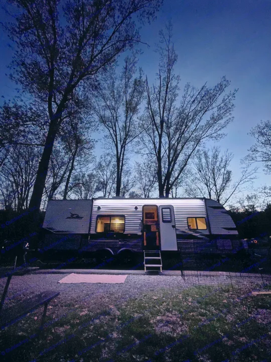 An RV rental glows at dusk, parked on a gravel campsite amidst silhouetted trees. Interior and exterior lights illuminate the peaceful scene.. Jayco Jay Flight 2024