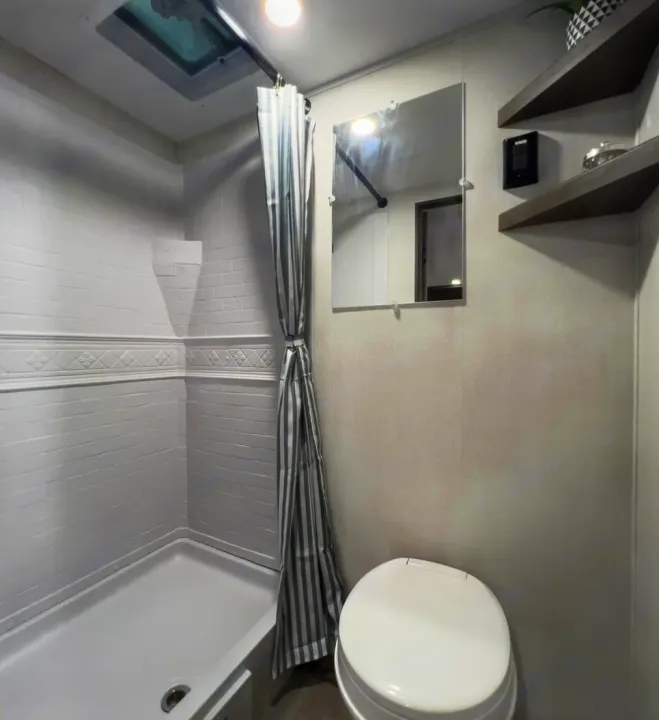 This RV rental's compact bathroom features a white shower, toilet, mirror, shelves, and a skylight.. Grand Design Transcend One 151BH 2025