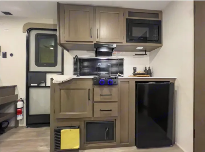 Inside an RV rental: a well-equipped kitchenette with microwave, stove, mini-fridge, and storage cabinets, ready for your travel.. Grand Design Transcend One 151BH 2025