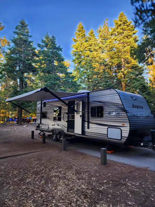 RV rental camping in a forest: A gray Jayco travel trailer is parked at a wooded campsite with its awning extended.. Jayco Jay Flight SLX Rocky Mountain Edition 2020