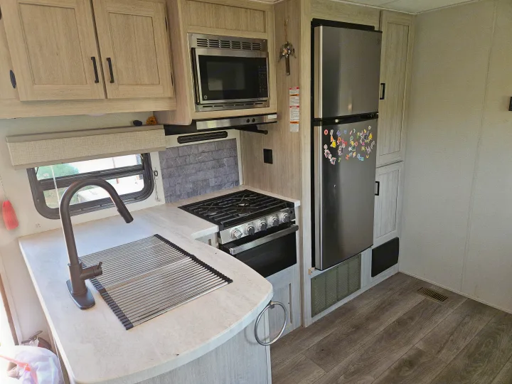 RV rental kitchen featuring a sink, stove, microwave, and refrigerator, with light wood cabinets and flooring.. Coachmen Catalina Legacy 2024