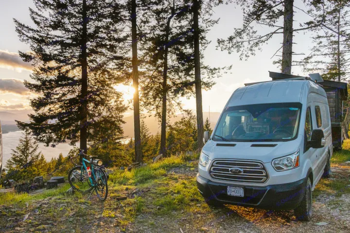 A white RV rental van at a scenic lakeside campsite, silhouetted by sunset over mountains. A bicycle rests nearby.. Ford Transit 2017