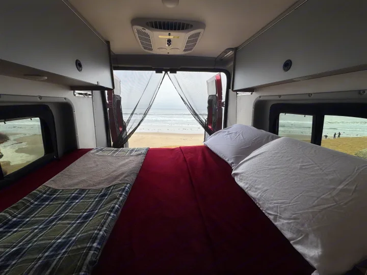 RV rental camper van interior. Features a cozy bed with blankets/pillows, and a clear beach/ocean view through open rear doors.. Winnebago Winnebago 2023