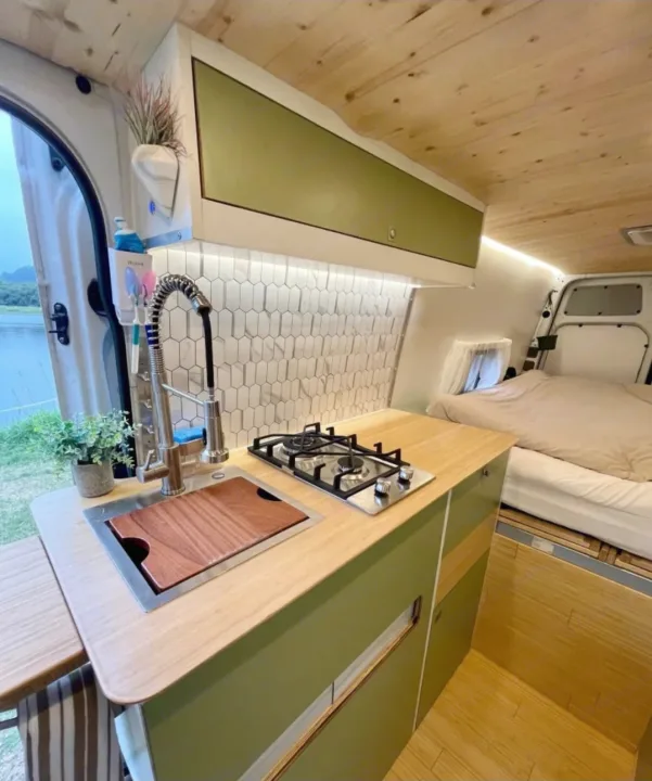Spacious RV rental interior with modern kitchen (sink, stove, tiled backsplash) and comfortable bed area by the window.. Mercedes Sprinter 2012