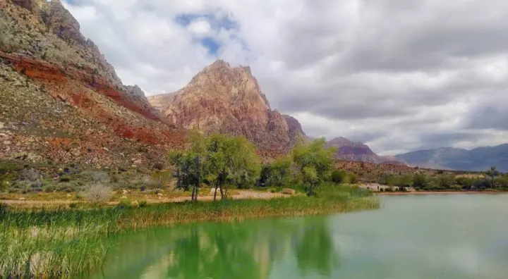 Serene green lake with tall reeds, flanked by dramatic red rock mountains under a cloudy sky. Perfect RV rental adventure.. Grand Design Transcend One 151BH 2025