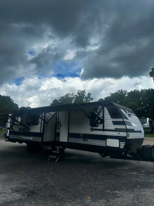 A Zinger travel trailer RV rental, with its awning out, parked on gravel under a dramatic cloudy sky.. Crossroads RV Zinger 2020