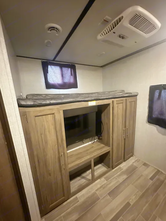 RV rental interior: bunk beds over a TV entertainment unit with storage, featuring a ceiling AC unit and wood-look plank flooring.. Crossroads RV Zinger 2020