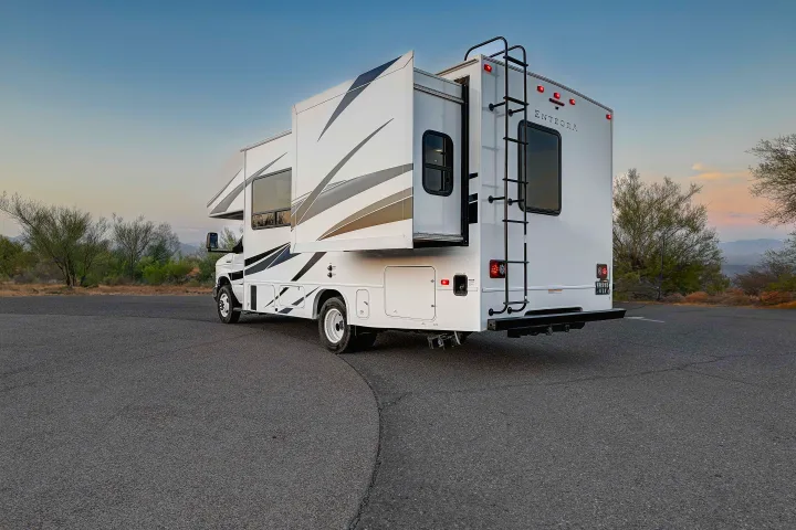 A white motorhome RV rental with extended slide-outs parked on asphalt, set against a desert landscape at dusk. Ideal for travel.. Entegra Coach Odyssey SE 2024