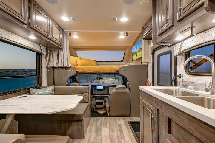 View inside an rv rental: dining booth, overhead bunk with yellow bedding, driver's cab, and kitchen with double sink.. Entegra Coach Odyssey SE 2024