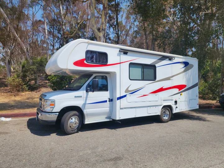 Bright white RV rental, with red and blue stripes, parked outdoors on a sunny day with tall trees. Ideal for camper getaways.. Forest River Forester 2018