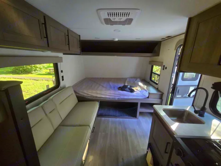 This RV rental interior features a comfortable bed, long sofa, and a kitchen with a sink. Overhead cabinets offer storage.. Grand Design 151BH 2025