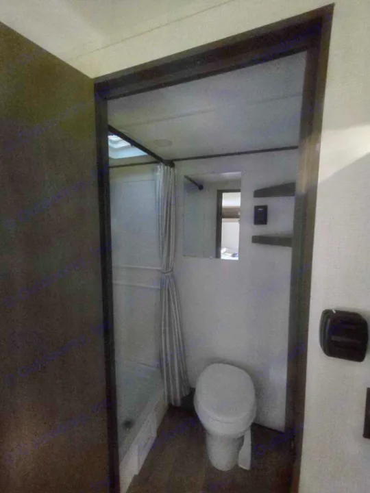 Inside an RV rental bathroom: a clean white toilet, shower with striped curtain, and wall shelves are visible.. Grand Design 151BH 2025