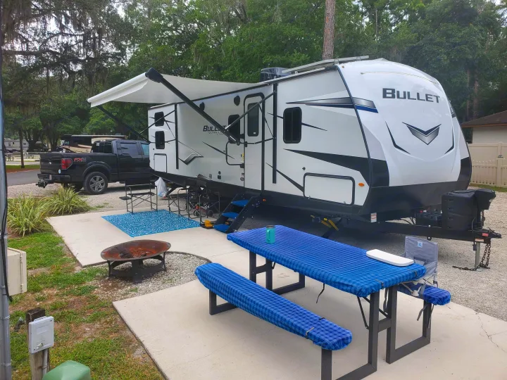 Comfy rv rental travel trailer with an extended awning, blue checkered picnic table, fire pit, and outdoor seating at a shady campsite.. Keystone RV Bullet Ultra Lite 2022