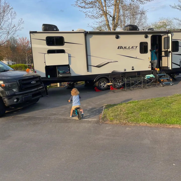 An RV rental adventure! A child on a balance bike, a large travel trailer, and a pickup truck parked at a scenic campground.. Keystone RV Bullet Ultra Lite 2022