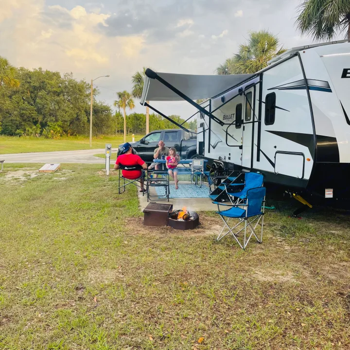 Family enjoying an **RV rental** at a campsite. A travel trailer with an extended awning, picnic table, and crackling campfire.. Keystone RV Bullet Ultra Lite 2022