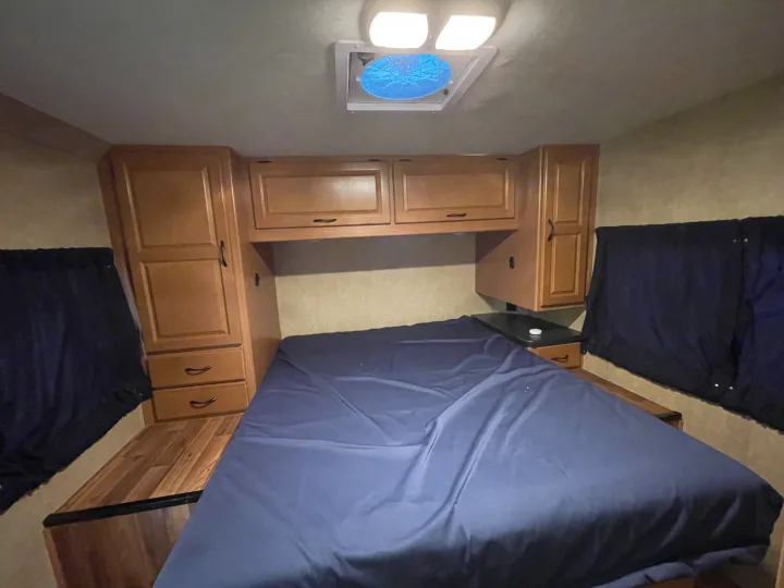 RV rental's cozy bedroom features a blue bed, wooden cabinets, and black-out window coverings. Functional camper interior.. Other Other 2017