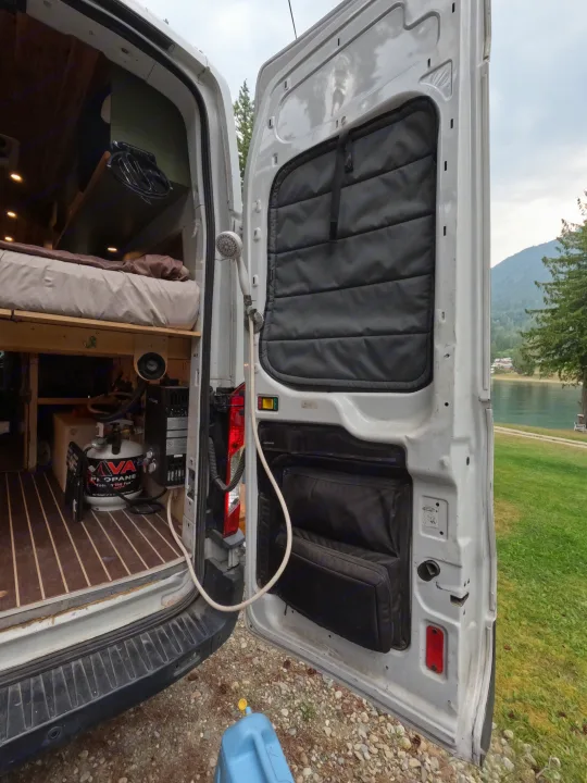 RV rental: Rear view inside a camper van, showing a bed, portable propane water heater, and outdoor shower with a lake backdrop.. Ford Transit 2017