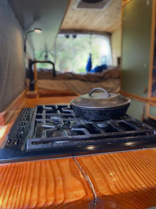 Cozy RV rental interior: Cast iron pot on a gas stove, with a bed and green outdoor view in the background.. Ford Transit 2017