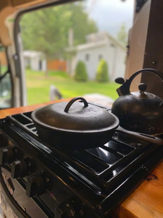 RV rental stovetop with a Lodge cast iron pot & kettle, overlooking a serene view of a house & green yard.. Ford Transit 2017