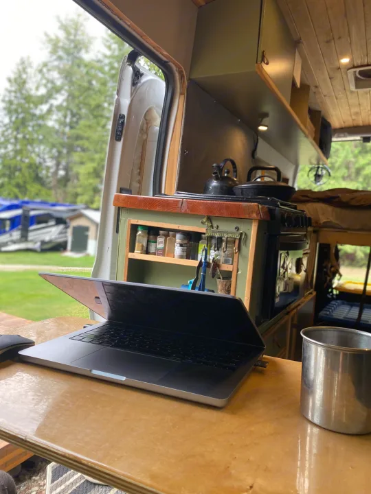 Work from anywhere in this cozy RV rental! A laptop and metal cup sit on the counter inside a camper van with its door open to nature.. Ford Transit 2017