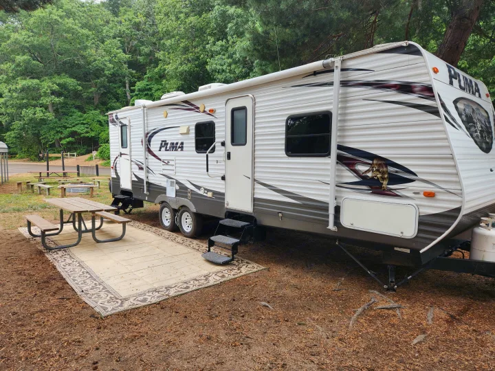 RV rental at a wooded campsite featuring a white travel trailer with an awning, picnic table on a rug, and green trees.. Forest River Palomino Puma 2013