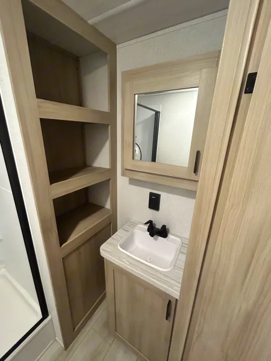 Clean RV rental bathroom with wood-toned vanity, white sink, black faucet, mirrored cabinet, and open shelving.. East to West Della Terra 2024