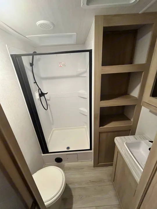 RV rental bathroom interior: compact shower, toilet, sink vanity with shelving, and light wood-look floor.. East to West Della Terra 2024