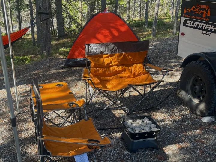 An RV rental campsite in a forest featuring an orange tent, two camping chairs, a portable fire pit, and a hammock.. Sunset park rv Sunray 109 2025