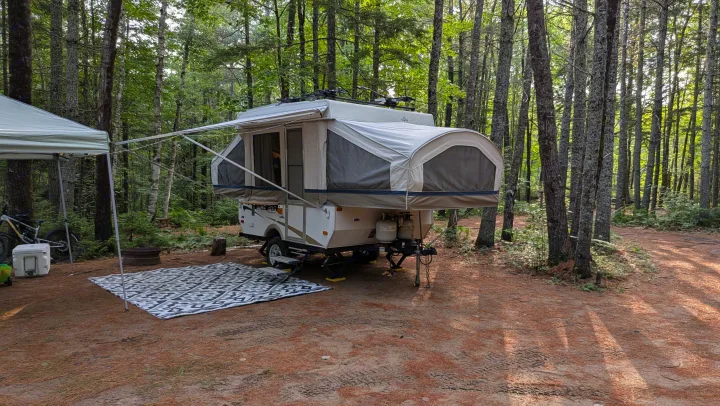 Pop-up RV rental fully set up in a sunlit forest campsite with an awning, outdoor rug, and nearby tent. Ideal for camping.. Coachmen Clipper 2008