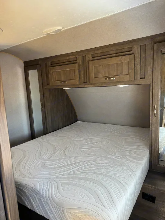 Inside an RV rental: a clean, inviting bedroom with a white patterned mattress, overhead cabinets, and wood accents.. Forest River vibe 2020