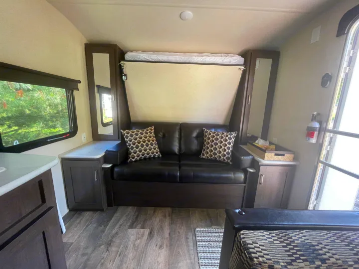 RV rental interior: compact living space with a black sofa, wall-mounted murphy bed (up), and storage cabinets.. Forest River Wildwood X-Lite 2018