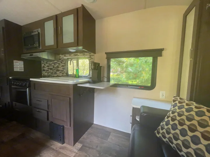 Modern RV rental kitchen & seating area with dark cabinets, stove, sink, and coffee maker. Window overlooks nature.. Forest River Wildwood X-Lite 2018