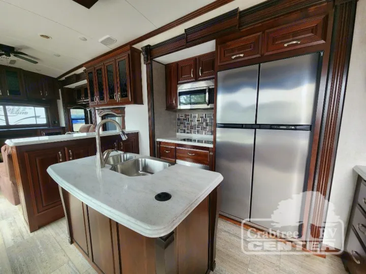 Spacious, modern RV kitchen interior featuring a large stainless steel refrigerator, island sink, and dark wood cabinetry. Ideal for your next RV rental.. Jayco Designer 39RE 2017