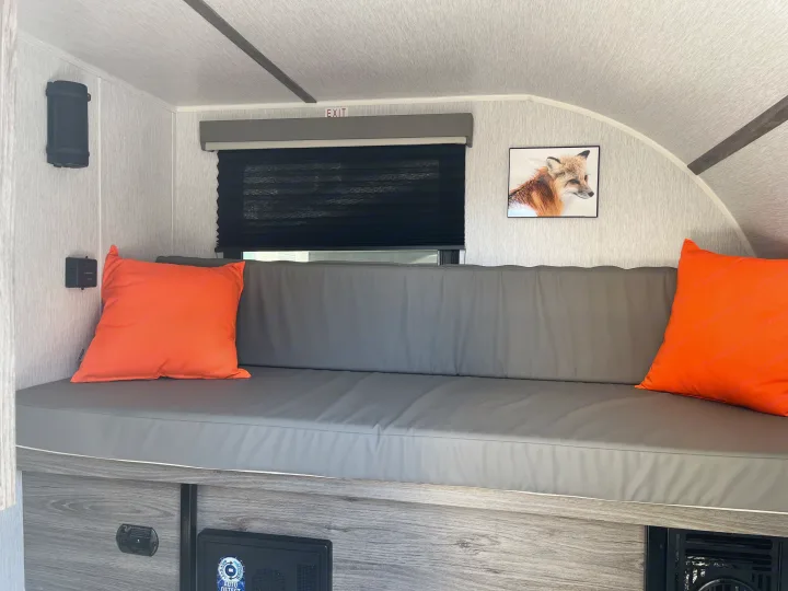 RV rental interior with a grey bench, bright orange pillows, black window blinds, fox art, and an EXIT sign.. Sunset Park RV SunRay 2025