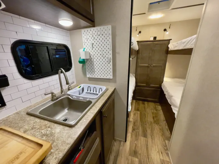 Inside an RV rental, view of the kitchen area with a sink, window, and pegboard, extending to bunk beds and a cabinet.. Sunset Park RV Sun Lite 2019