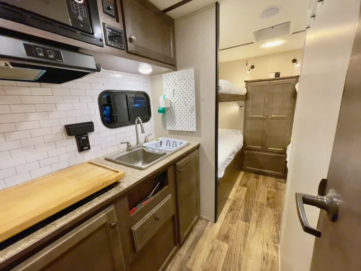 Inside this RV rental: functional kitchen with sink, microwave, tiled backsplash, & comfy bunk beds. Ideal for travel adventures!. Sunset Park RV Sun Lite 2019