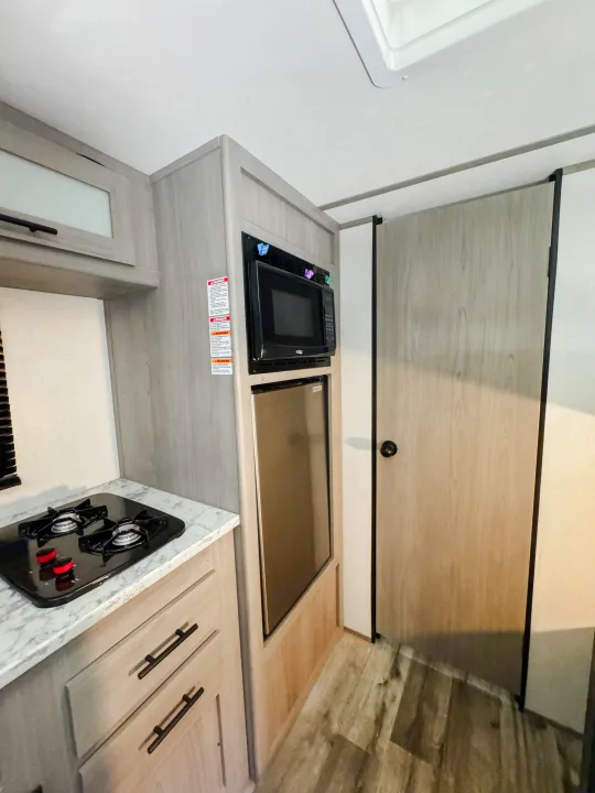 This RV rental kitchen features a compact two-burner stove, microwave, fridge, and light wood cabinets.. Dutchmen Coleman Lantern LT 2023