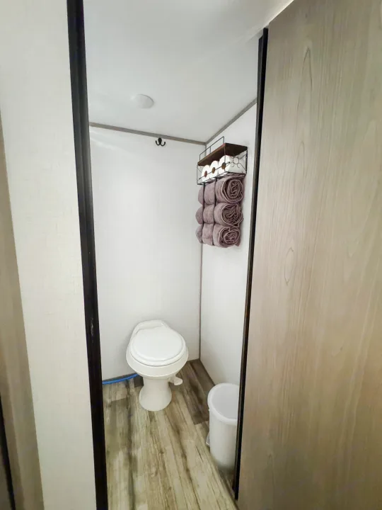 Compact RV rental bathroom: white toilet, wall storage for towels/TP, and light wood floor. Organized & functional.. Dutchmen Coleman Lantern LT 2023