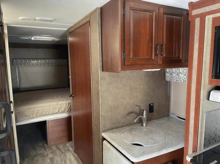 Compact RV rental interior featuring a cozy bedroom with a bed, adjacent to a compact kitchen area with a sink and overhead cabinets.. Forest River Sunseeker 2016