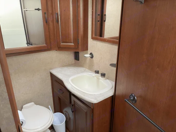 Compact RV rental bathroom with a white toilet, sink, and wooden vanity, featuring a wall-mounted mirror.. Forest River Sunseeker 2016