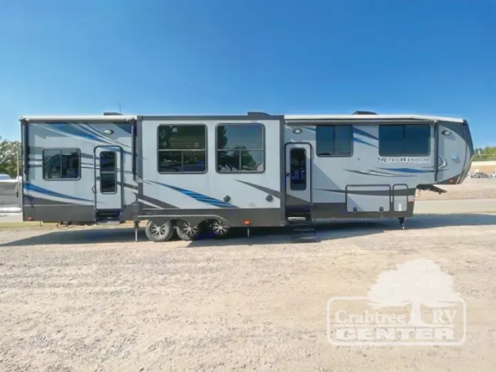 A long, gray 'Road Warrior' fifth-wheel RV with multiple windows and doors, parked on a gravel lot. Consider for RV rental.. Heartland Road Warrior 425 2016