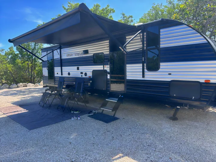 Spacious Grey Wolf RV rental with extended awning, chairs, and mat at a sunny gravel campsite for outdoor relaxation.. Forest River Cherokee 2022