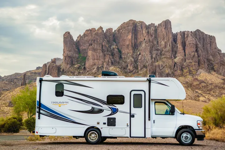 White Triumph RV rental parked on a scenic road, with dramatic desert mountains towering in the background.. Nexus Other 2026